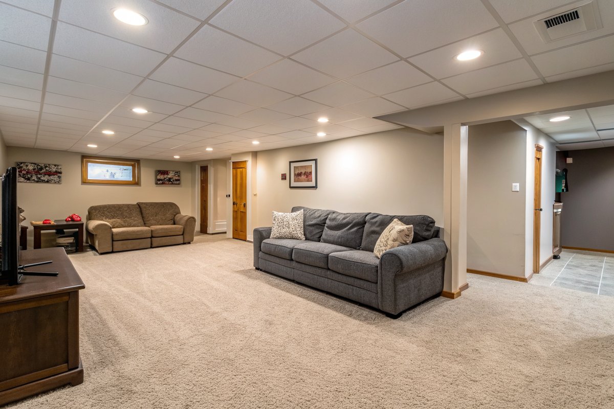 A dry finished basement family room with light carpet, a gray sectional and a small egress window